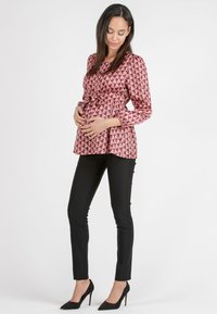 Gravidblus i rosa med geometriska mönster, peplumdesign knyten i midjan; ihopparat med svarta tajta byxor och högklackade skor.