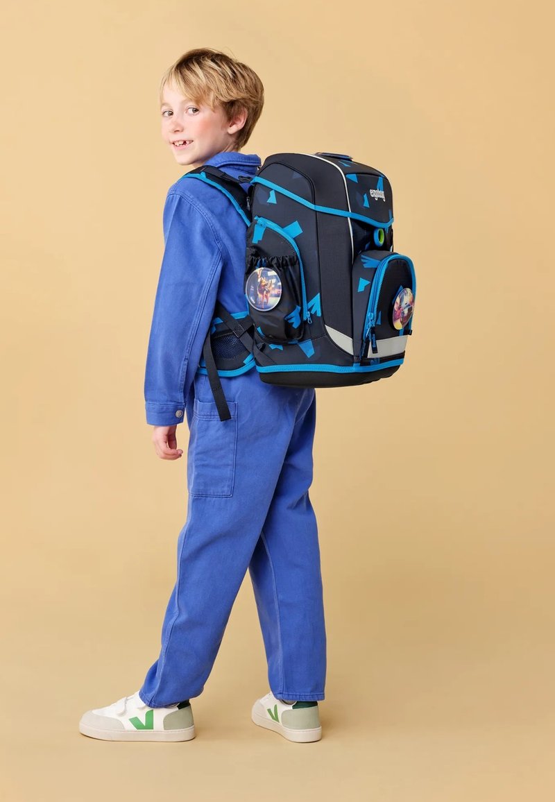 Sac à dos noir avec des accents bleus, plusieurs poches et un graphisme circulaire. Enfant en tenue bleue se tient devant un fond beige.