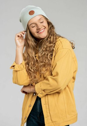 Jasnoniebieska dziana czapka z brązową skórzaną łatą, noszona z żółtą kurtką dżinsową. Modelka ma długie, kręcone włosy i miękkie tło.