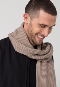 Scialle in maglia a coste beige drappeggiato attorno al collo, caratterizzato da una consistenza morbida e un aspetto caldo, abbinato a un indumento nero sotto.