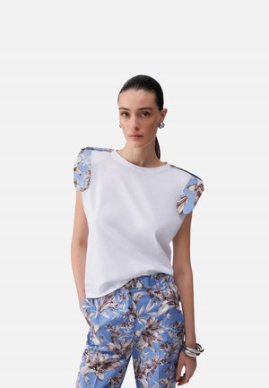 Donna con capelli scuri raccolti indossa una canotta bianca con spalline floreali blu e pantaloni abbinati a fiori blu, con orecchini e braccialetti d'argento.