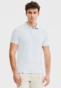 Jasnoniebieska koszulka polo wykonana z miękkiego materiału, z klasycznym kołnierzykiem, krótkimi rękawami i zapięciem na trzy guziki, noszona z kremowymi spodniami.