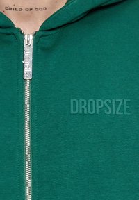 Grøn zip-up hoodie lavet af blødt stof, med en sølvfarvet lynlås, stort tekst "DROPSIZE" på forsiden og foret hætte.