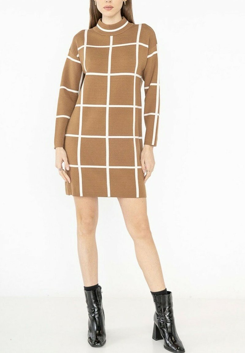 Robe tricotée marron avec un col haut, présentant des motifs de grille blancs. Manches longues et coupe droite, associée à des bottines à talons noires.