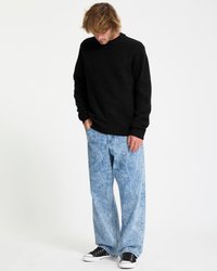 Man with tousled hair wearing a black sweater, loose light blue acid-wash jeans, and black sneakers looking downward with hands in pockets.