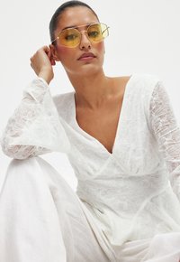 Haut blanc brodé avec des motifs floraux et des manches fluides, associé à des lunettes de soleil hexagonales teintées de jaune, sur fond clair.