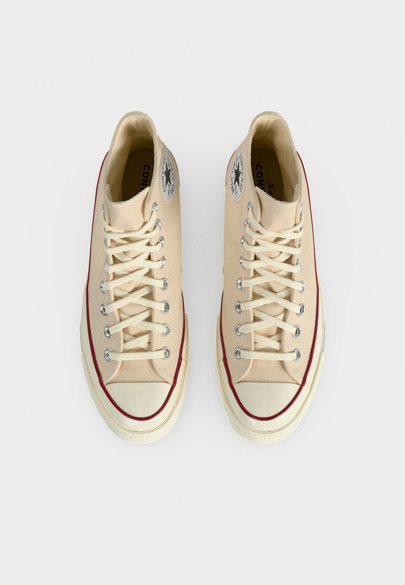 Ein Paar beige High-Top-Converse-Sneaker mit weißen Schnürsenkeln, Gummisohlen und rotem Streifendetail, von oben auf einfachem Hintergrund gesehen.