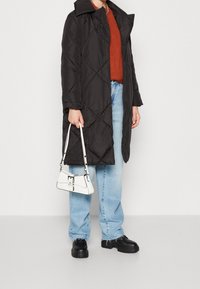 Osoba oblečená v černé prošívané bundě, oranžovém pleteném svetr, světle modrých džínách a černých robustních botách, držící malou bílou kabelku po boku.