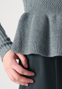 Gros plan sur une main reposant près de l'ourlet d'un pull gris côtelé et d'un pantalon en tissu foncé sur un fond uni.