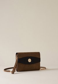 Sac en bandoulière texturé marron avec accent noir et fermeture circulaire dorée, doté d'une bandoulière assortie avec des sections de chaîne dorée, sur fond beige.