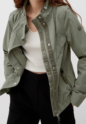 Kvinde iført en åben olivengrøn jakke over en hvid ribstrikket kort top og sorte bukser, med hænderne i jakkelommerne.