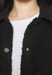 Svart texturerad jacka med en fluffig finish, med silverfärgade tryckknappar och krage, lagrad över en vit t-shirt.