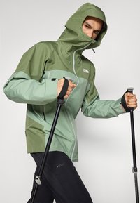 Veste en nylon verte avec capuche, présentant un design en blocs de couleurs vert clair et foncé, une fermeture éclair à l'avant et des poignets ajustables. Tenant des bâtons de trekking.