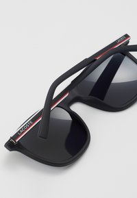 Ochelari de soare negri cu finisaj mat, lentile rectangulare, accente roșii și albe pe brațe și brandul Lacoste pe laterale.