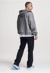 Grau melierter Kapuzenpullover mit lockerem Schnitt, gerippten Bündchen und Saum, kombiniert mit schwarzer Hose und blauen Sneakern, von hinten gesehen.