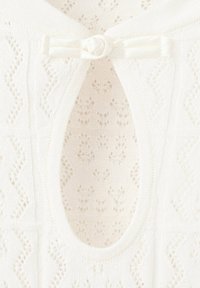Gros plan d’un tissu tricoté blanc avec un motif ajouré en losange et un petit bouton couvert de tissu avec une fermeture à boucle au-dessus d’une ouverture en forme de goutte.