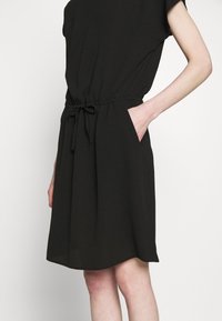 Vestido negro de longitud hasta la rodilla con mangas cortas y un lazo en la cintura, con la mano en el bolsillo lateral, usado por una persona de piel clara.