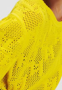 Suéter amarillo de punto con un patrón calado, diseño de zigzag texturizado y un corte holgado. La tela es ligera y transpirable.