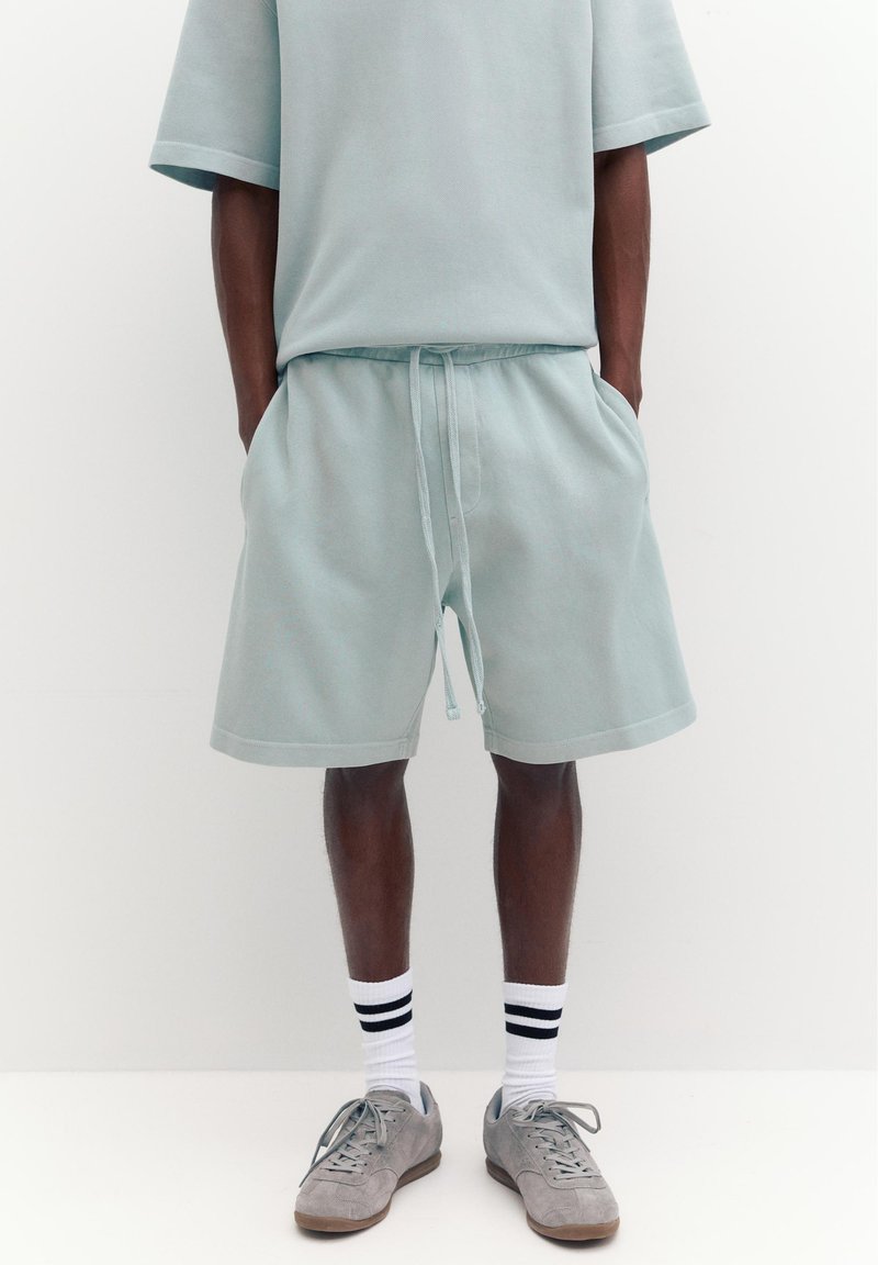 Homme portant un short gris clair avec un cordon, un haut à manches courtes assorti, des chaussettes blanches à rayures noires et des baskets grises, debout les mains dans les poches.