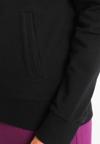 Sudadera negra con un bolsillo lateral, puños y dobladillo acanalados. La tela es suave y ligera, complementando una prenda morada debajo.