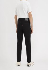 Jeans de mezclilla negra con un ajuste slim, bolsillos traseros y una cinturilla texturizada. Llevados con una camiseta blanca de manga corta y zapatos de color claro.