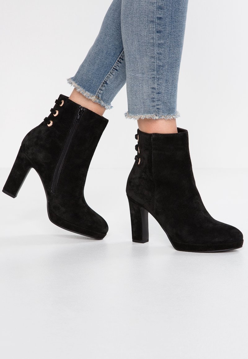 Bottines en suède noir montant à la cheville, à talons blocs et fermetures éclair latérales, portées avec un jean bleu raccourci à ourlet effiloché.