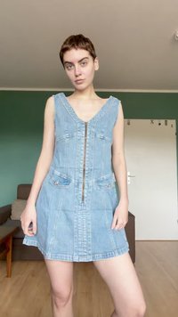 Personne aux cheveux courts portant une robe en denim bleu clair sans manches avec une fermeture éclair à l'avant, debout dans une pièce aux murs verts et au sol en bois.