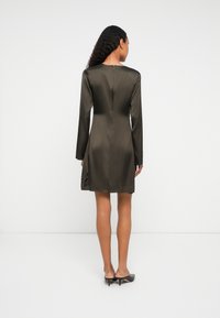 Sieviete ar gariem cirtainiem matiem ģērbta tumši brūnā satīna kleitā ar garām piedurknēm un melnām kaķu papēžiem, attēlota no aizmugures uz baltā fona.