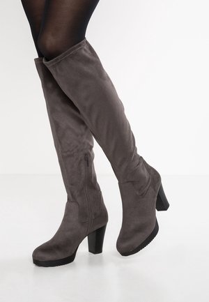 Høje grå ruskindsstøvler med hæl, båret over sorte strømpebukser, med sidelommer og chunky hæle på en hvid baggrund.