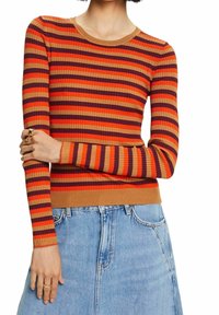 Femme portant un pull côtelé à manches longues avec des rayures horizontales orange, rouge et violettes, associé à un jean en denim bleu clair.