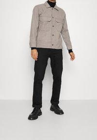 Man in een beige knoopjas met borstzakken, een zwarte coltrui, zwarte opgerolde broek en zwarte chunky laarzen op een witte achtergrond.