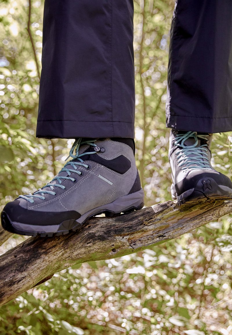 Bottes de randonnée grises avec des accents noirs, en suède texturé, lacets teal, et une semelle robuste, reposant sur une branche d'arbre dans un cadre naturel.