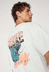 Hvid oversized T-shirt med et grafisk tryk på ryggen med "NIKE" i sort og ferskenfarvet, samt et blåt design med teksten "BASKETBALL."