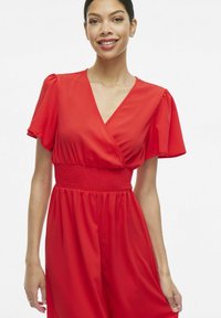 Červené šaty s výstrihom do V a krátkymi púzdrovými rukávmi. Obsahujú skladaný pás a splývavú sukňu. Hladká textúra látky.