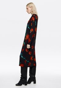 Blondynka w profilu, ubrana w długą czarną suknię w czerwone kwiatowe wzory oraz czarne botki na obcasie na białym tle.