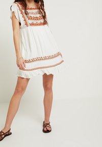 Valkoinen mekko lyhyillä hihoilla, oranssia ja ruskeaa brodeerausta sekä rummunreunaa; kevyt kangas, joka laskeutuu kauniisti.