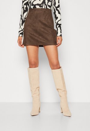 Kvinde iført en brun ruskind mini nederdel, sort- og hvidmønstret langærmet top og beige knæhøje støvler med hæl, stående mod en hvid baggrund.