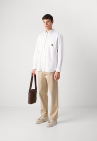 Man in een witte overhemd, beige broek, crèmekleurige sneakers, met een bruine schoudertas tegen een plain witte achtergrond.