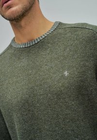 Zielony sweter dziany z okrągłym dekoltem. Posiada teksturowane wykończenie i kontrastujący szary ściągacz przy kołnierzyku. Mały logo na piersi.