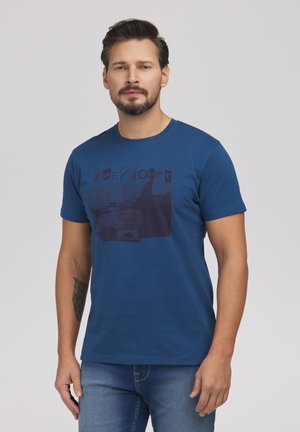 Mężczyzna z brodą i tatuażem, ubrany w niebieski t-shirt z grafiką i dżinsy, stojący na jednolitym szarym tle.