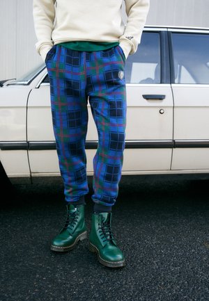Person trägt blau-grüne karierten Jogginghose und grüne Schnürstiefel, steht vor einem weißen Oldtimer auf schwarzem Asphalt.