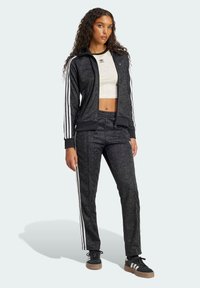 Čierny športový teplákový komplet s bielymi pruhmi, kvetinová textura na látke, zapínacia bunda, priliehavý crop top a zodpovedajúce nohavice. Čierne tenisky.