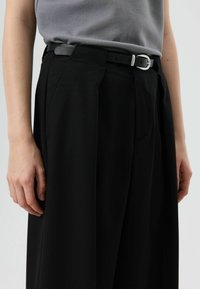 Zwarte broek met hoge taille en wijde pijpen, voorzien van plooien aan de voorkant en een aangesloten taille. Gecombineerd met een grijs aansluitend topje en een zwarte riem met zilveren gesp.