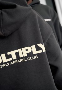Sudadera negra con gran texto blanco "MULTIPLY APPAREL CLUB" impreso en la parte posterior, con una textura suave y un diseño estándar de capucha.