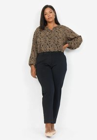 Schwarze und goldfarbene gemusterte Bluse mit V-Ausschnitt, luftigen Ärmeln, kombiniert mit taillierten schwarzen Hosen und nude-farbenen Ballerinas. Glatter Stoff und figurbetonte Passform.