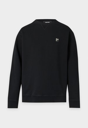 Jeune homme portant un sweat-shirt noir et un short noir avec texte blanc, chaussettes blanches et baskets noires, debout devant un fond uni.