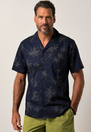 Man met baard, gekleed in een donkerblauw overhemd met korte mouwen met palmbomenpatroon en lichtgroene korte broek, staand met één hand in de zak.