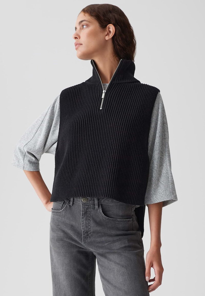 Frau in einem schwarzen, gerippten, ärmellosen Strickpullover mit einem halben Reißverschluss über einem grauen 3/4-Ärmel-Oberteil und dunkelgrauen Jeans, die zur Seite schaut.