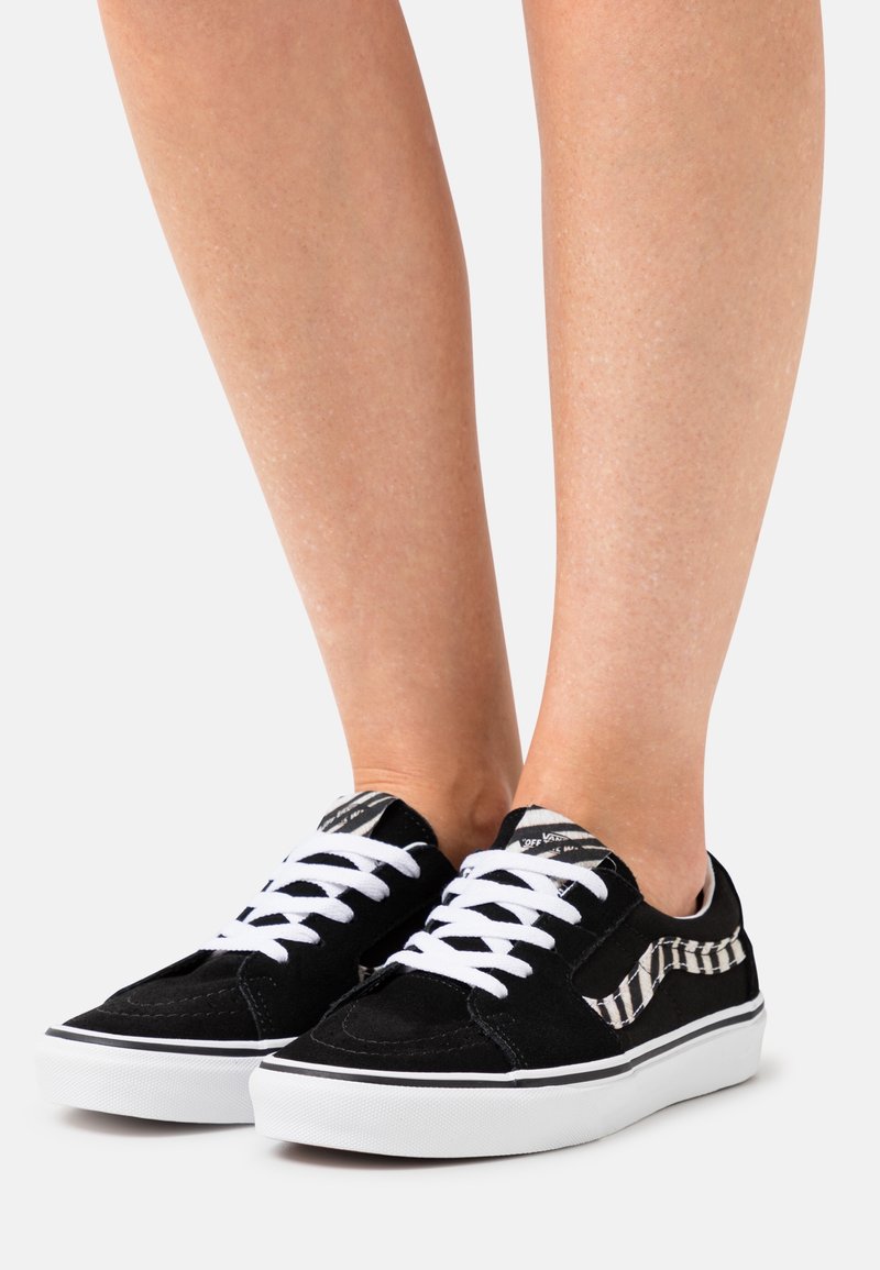 Jambes portant des baskets basses noires avec des lacets blancs, des semelles blanches et une bande latérale rayée noir et blanc sur un fond uni.