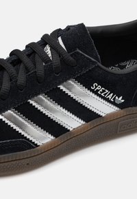 Czarny zamszowy sneaker z trzema metalicznymi srebrnymi paskami, teksturowaną gumową podeszwą i czarnymi sznurówkami. Logo "SPEZIAL" na bocznej części.